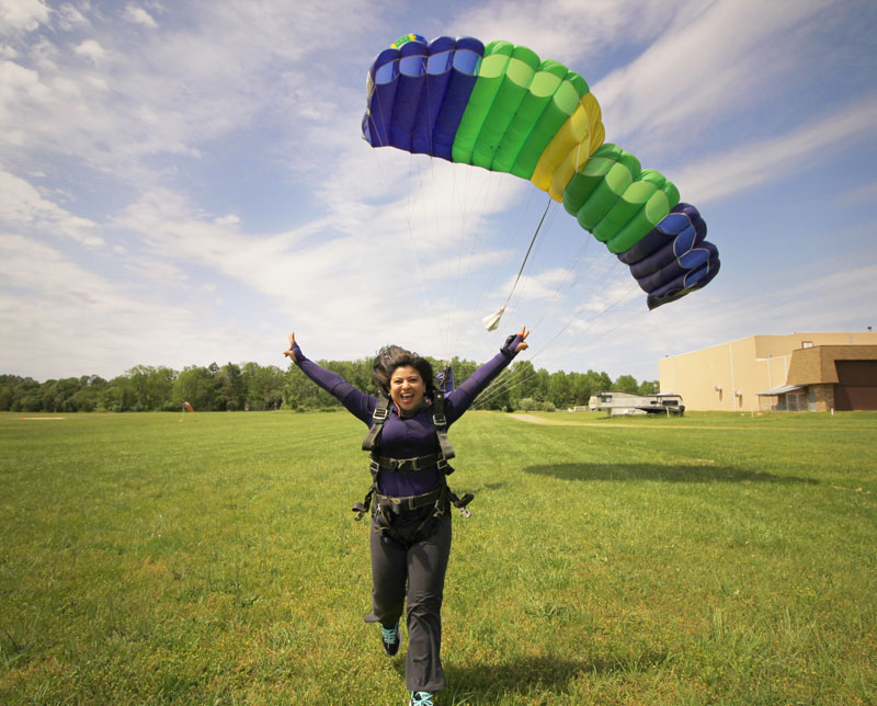Smiling skydiver landing in grassy field with canopy floating behind her.