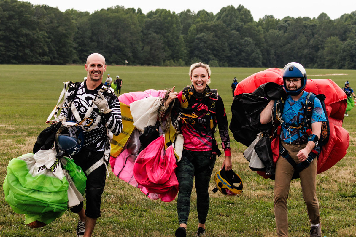 Three skydivers smiling on the ground with their harnesses on carrying deflated canopies after landing from a skydive.