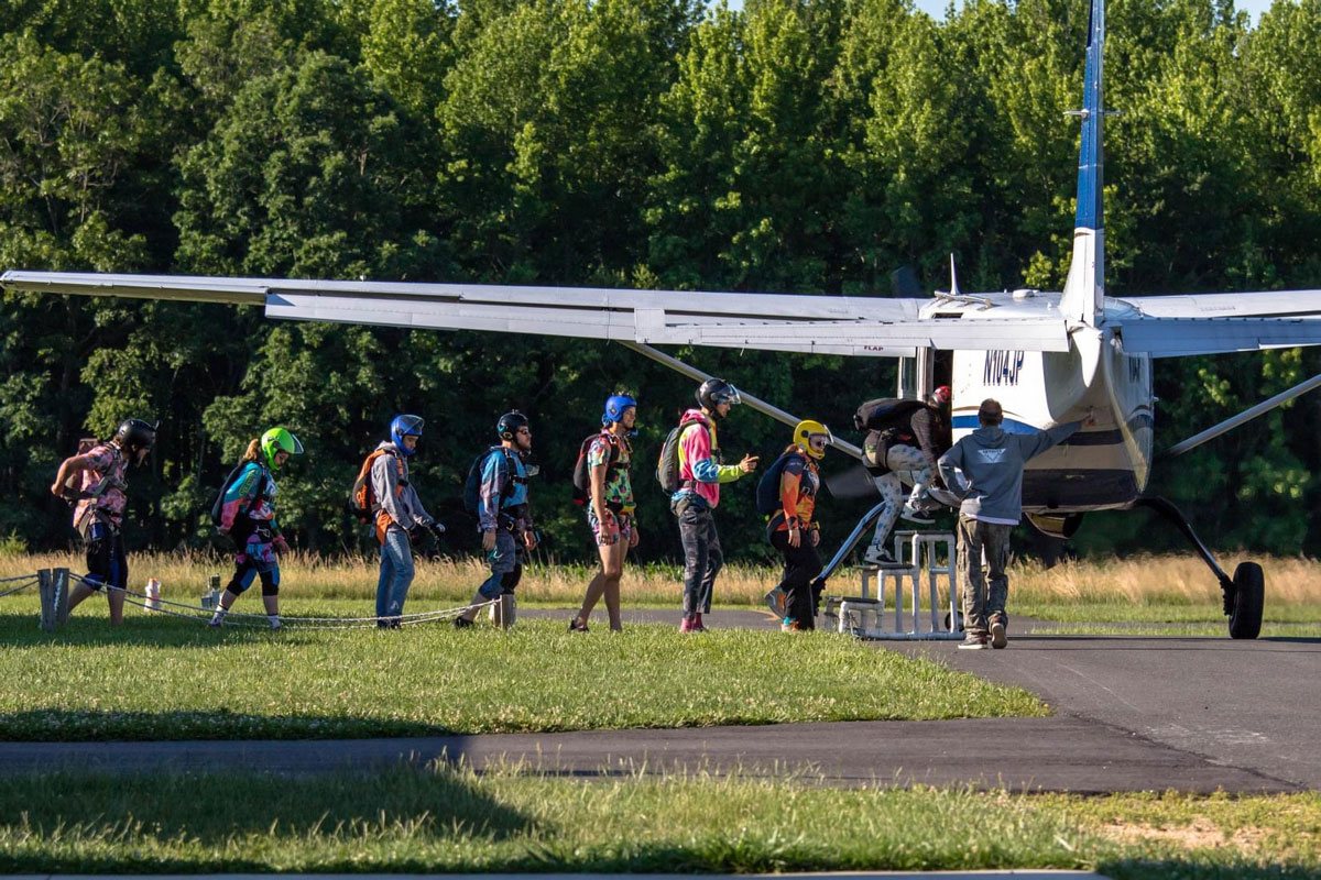 A line of skydivers in their harnesses with heltmets on, walking through a sunny field heading and boarding the plane on the airstrip.
